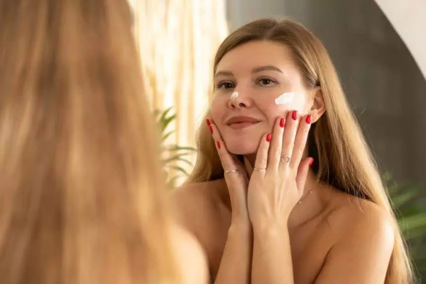 An young woman applies a moisturizing anti-aging cream to her face in front of a bathroom mirror.