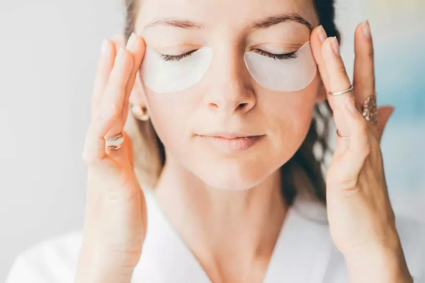 Attractive mature woman in white bathrobe puts anti aging eye patch on face taking care of skin