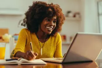 African American woman is smiling at the laptop and writing down in the textbook.