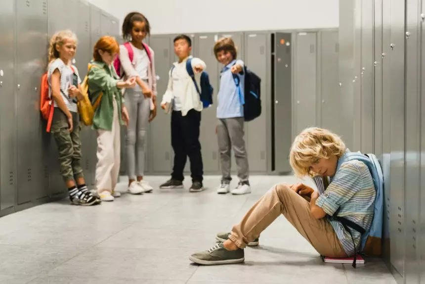 Sad crying schoolboy pupil sitting on the floor at the school hall while his classmates teenagers